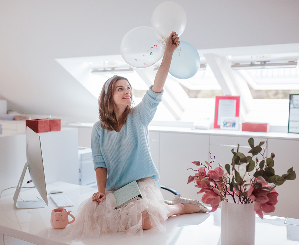 Mag. Dominika Schweiger-Kwiatkowski sitzt auf ihrem Schreibtisch und hält drei Luftballons in der Hand.